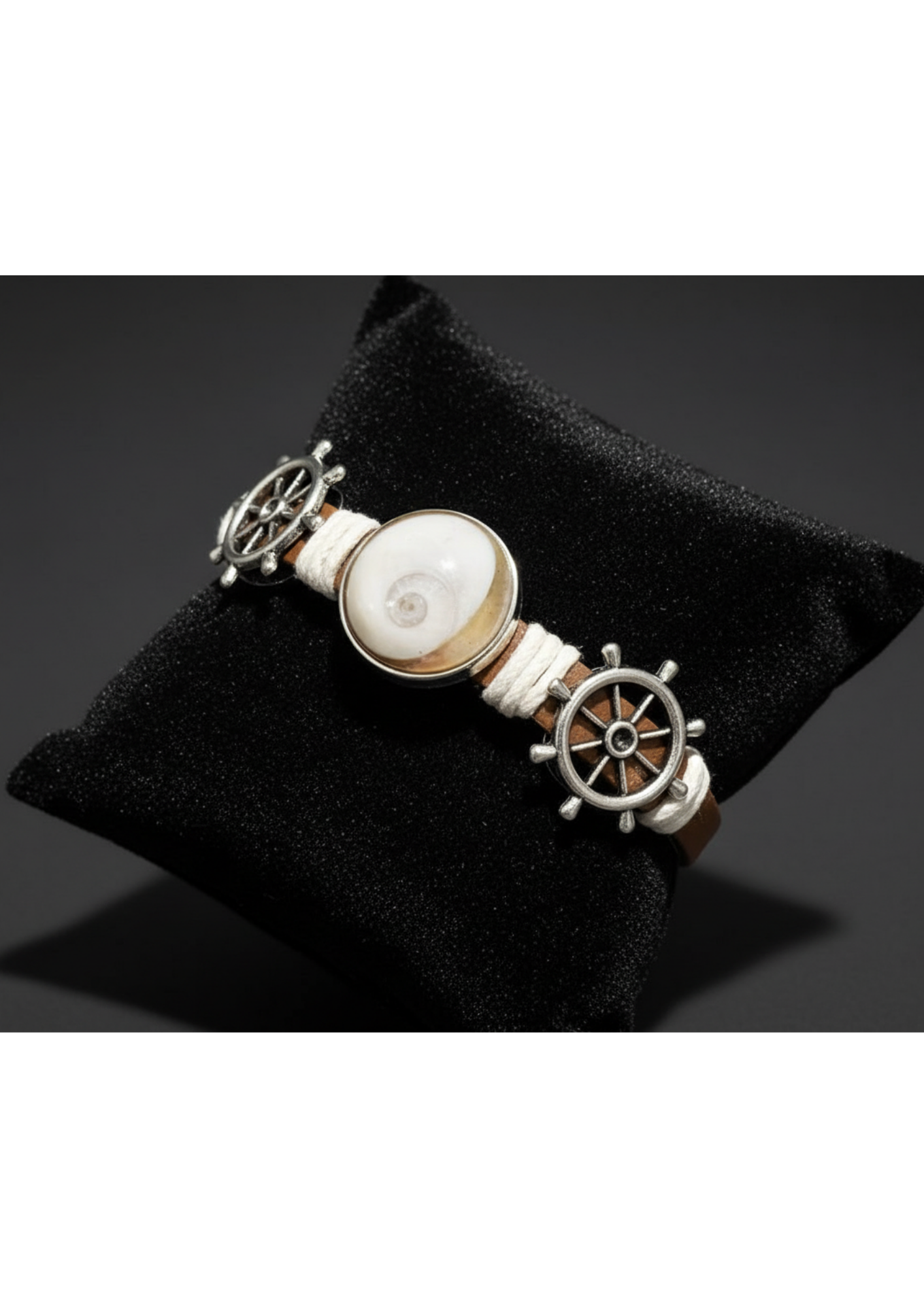 Bracelet homme œil de Sainte‑Lucie sur cuir marron, cordons écrus, décoré de deux roues de gouvernail métalliques, présenté sur coussin noir, style marin.