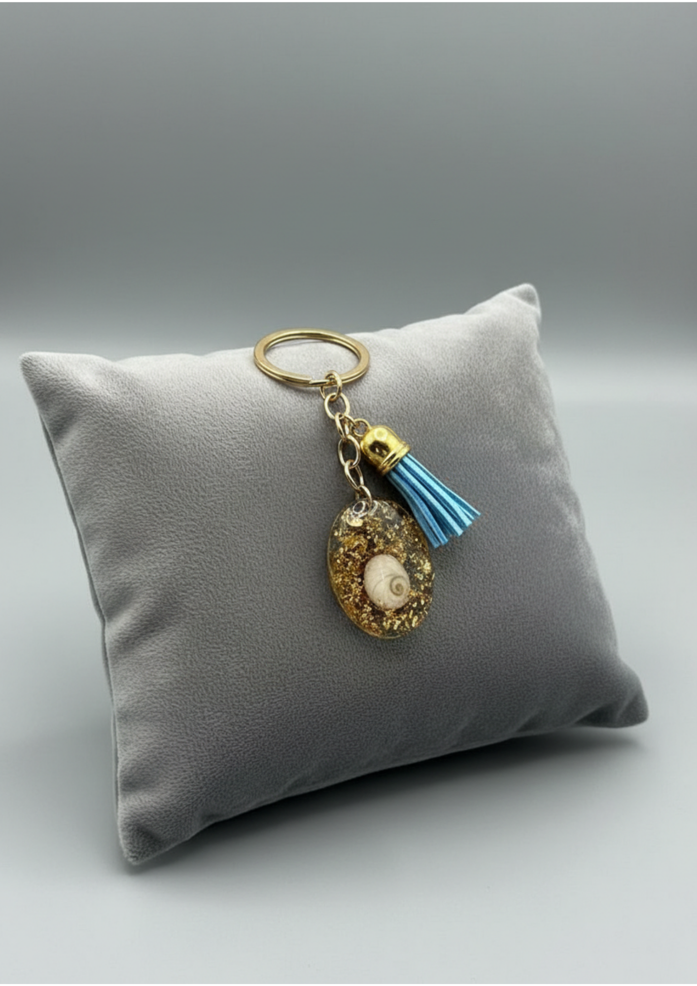 Porte-clés avec médaillon Œil de Sainte‑Lucie inclus dans résine dorée, anneau métal or et pampille bleue, présenté sur coussin gris, style marin chic.