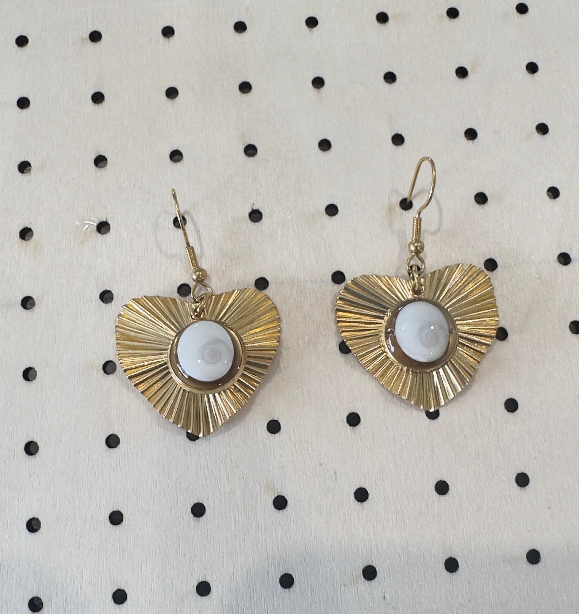 Vue rapprochée des boucles d'oreilles Astra dorées, pendentif cœur à rayons et cabochon Oeil de Sainte-Lucie