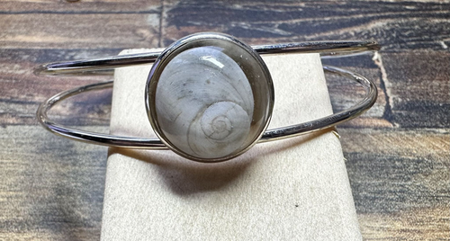 Bracelet jonc argenté avec médaillon rond serti d’un Œil de Sainte‑Lucie gris, posé sur boîte beige, fond bois, style minimaliste marin.