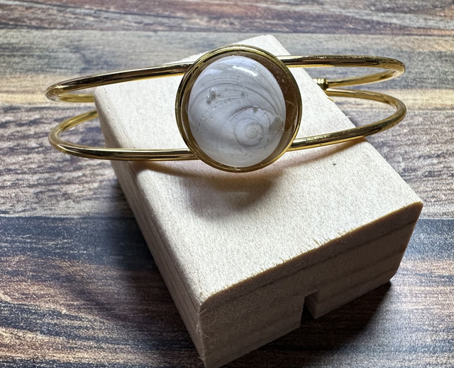 Bracelet jonc doré avec médaillon rond serti d’un Œil de Sainte‑Lucie gris, posé sur boîte beige, fond bois, style minimaliste marin.