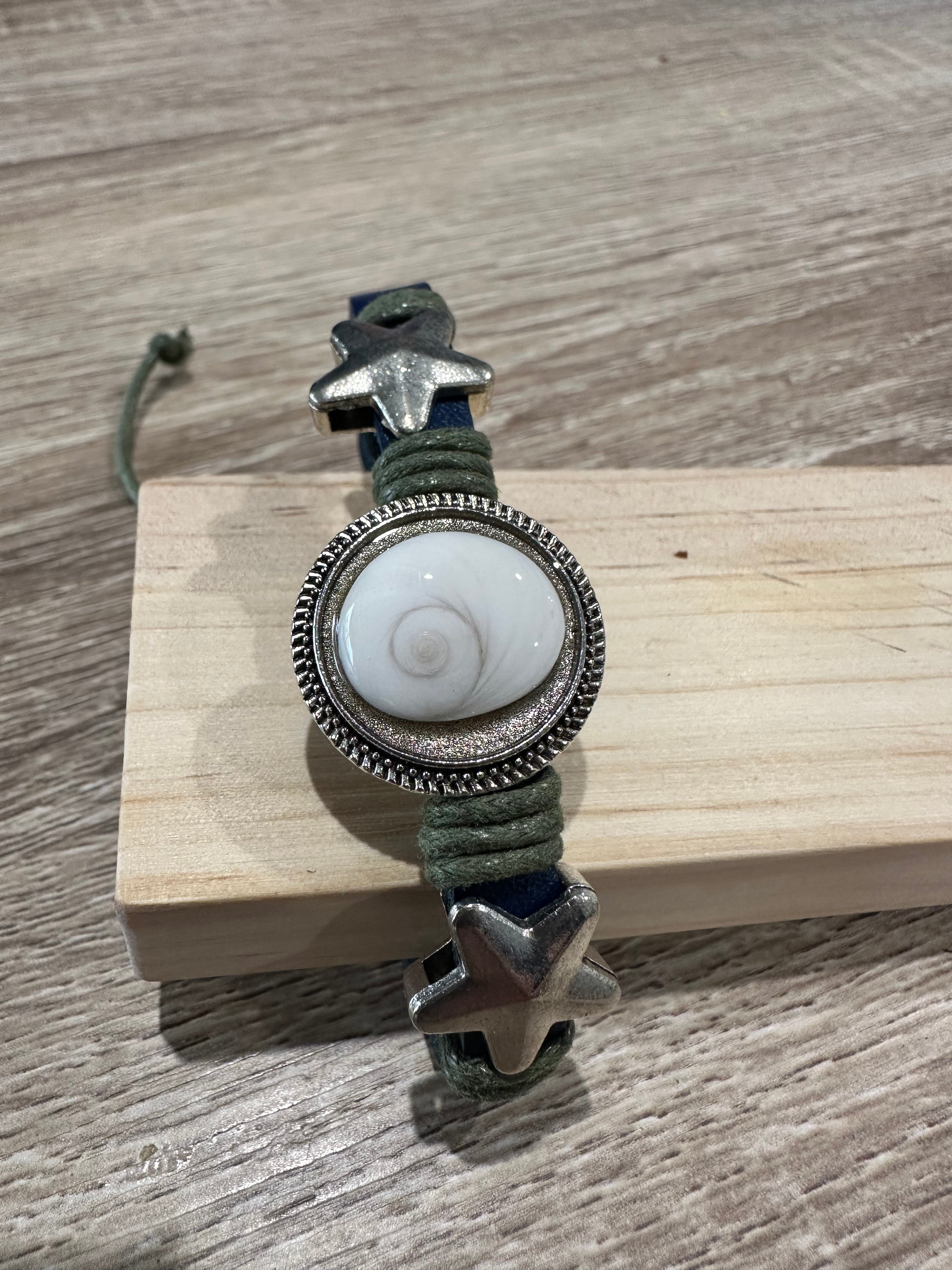 Bracelet ajustable en cuir bleu marine et cordon vert kaki tressé, avec deux breloques en forme d'étoile de mer argentée de chaque côté d'un grand cabochon rond serti en métal ciselé, contenant un œil de Sainte-Lucie (coquillage spiralé blanc nacré). Présenté sur un support en bois clair.