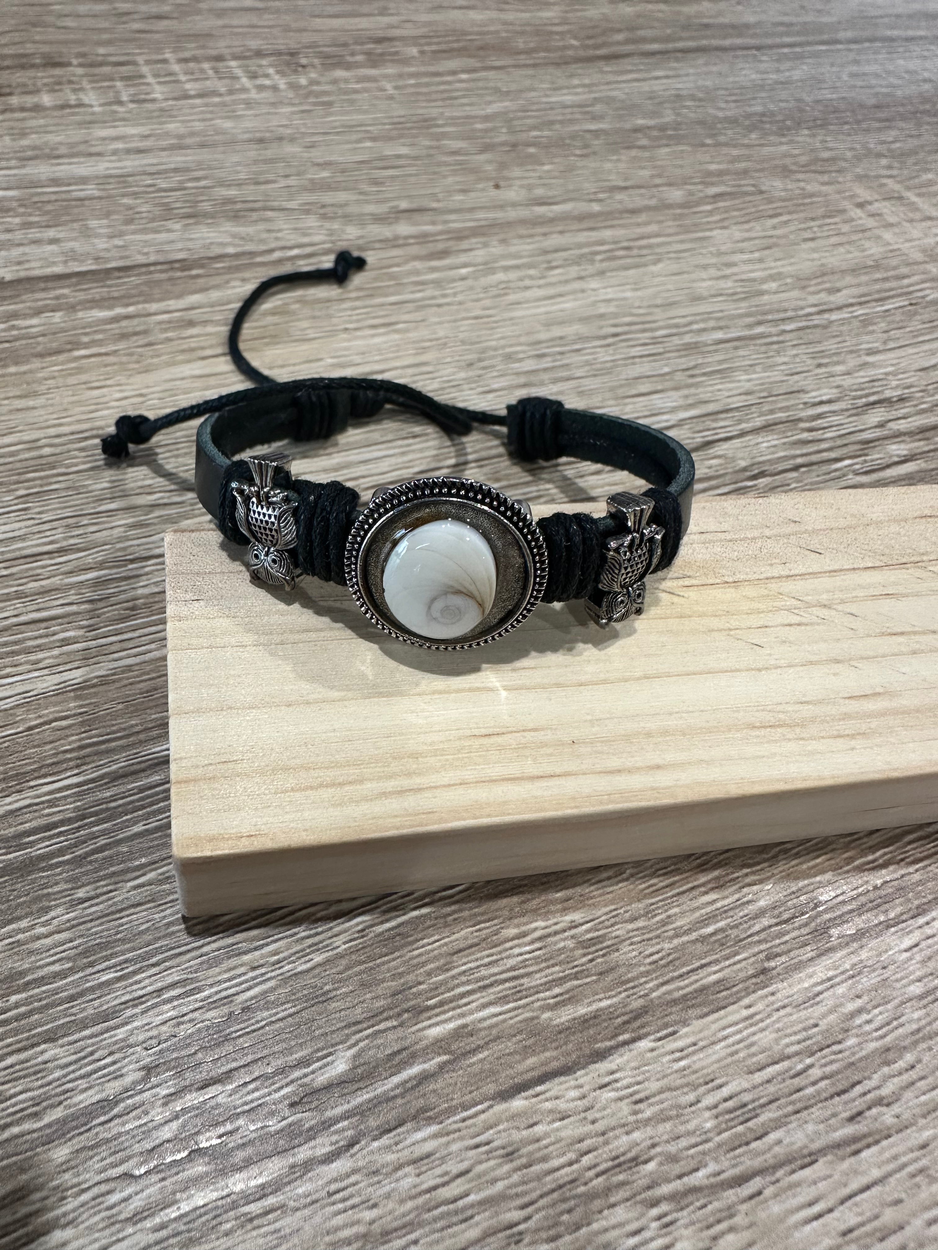 Bracelet ajustable à cordon noir tressé, orné de deux breloques en forme de hibou argenté de chaque côté d'un cabochon rond serti en métal argenté ciselé, contenant un œil de Sainte-Lucie (coquillage spiralé blanc nacré). Présenté sur un support en bois clair.