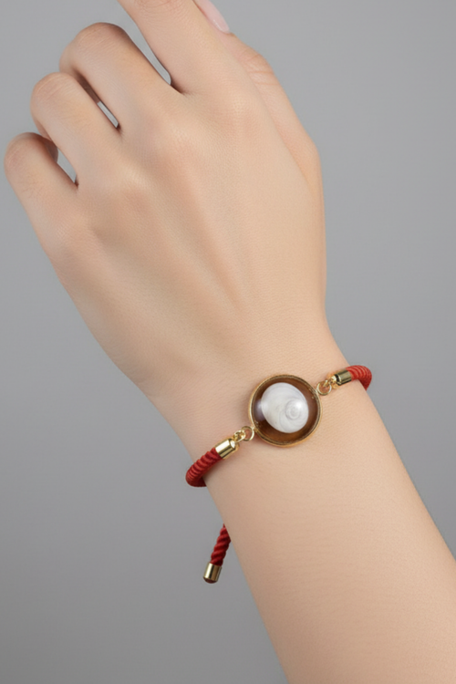 Bracelet femme cordon rouge au poignet, médaillon rond doré serti d’un Œil de Sainte‑Lucie blanc, embouts dorés, style chic et marin sur fond gris.