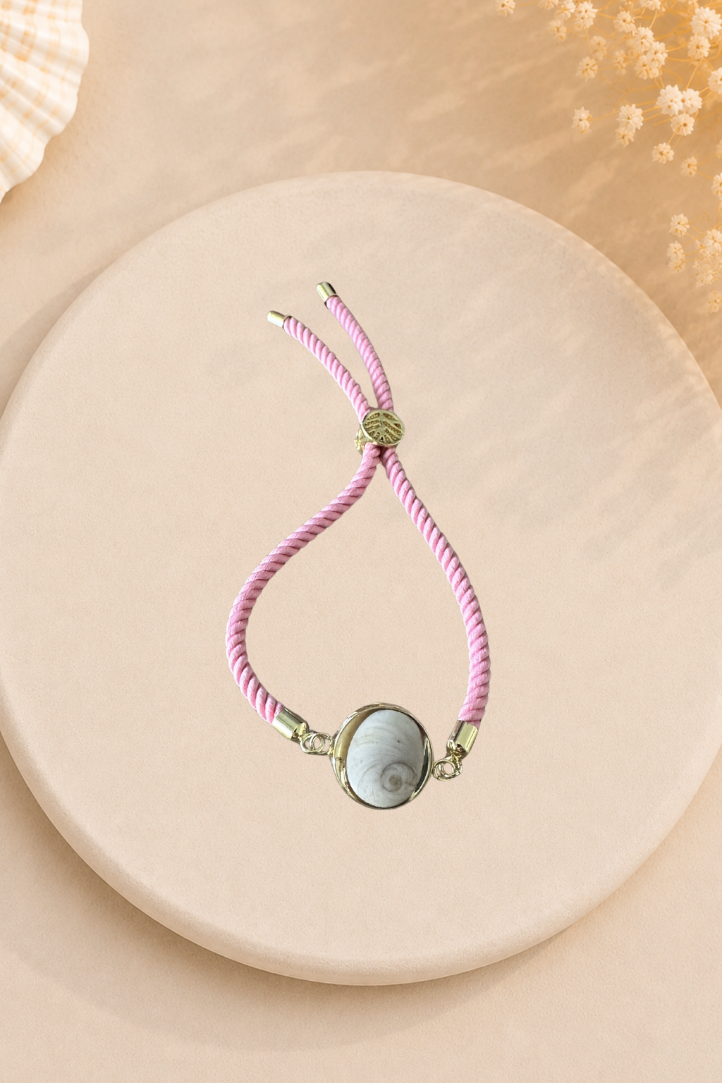 Bracelet femme cordon rose au poignet, médaillon rond doré serti d’un Œil de Sainte‑Lucie blanc, embouts dorés, style chic et marin sur fond gris.