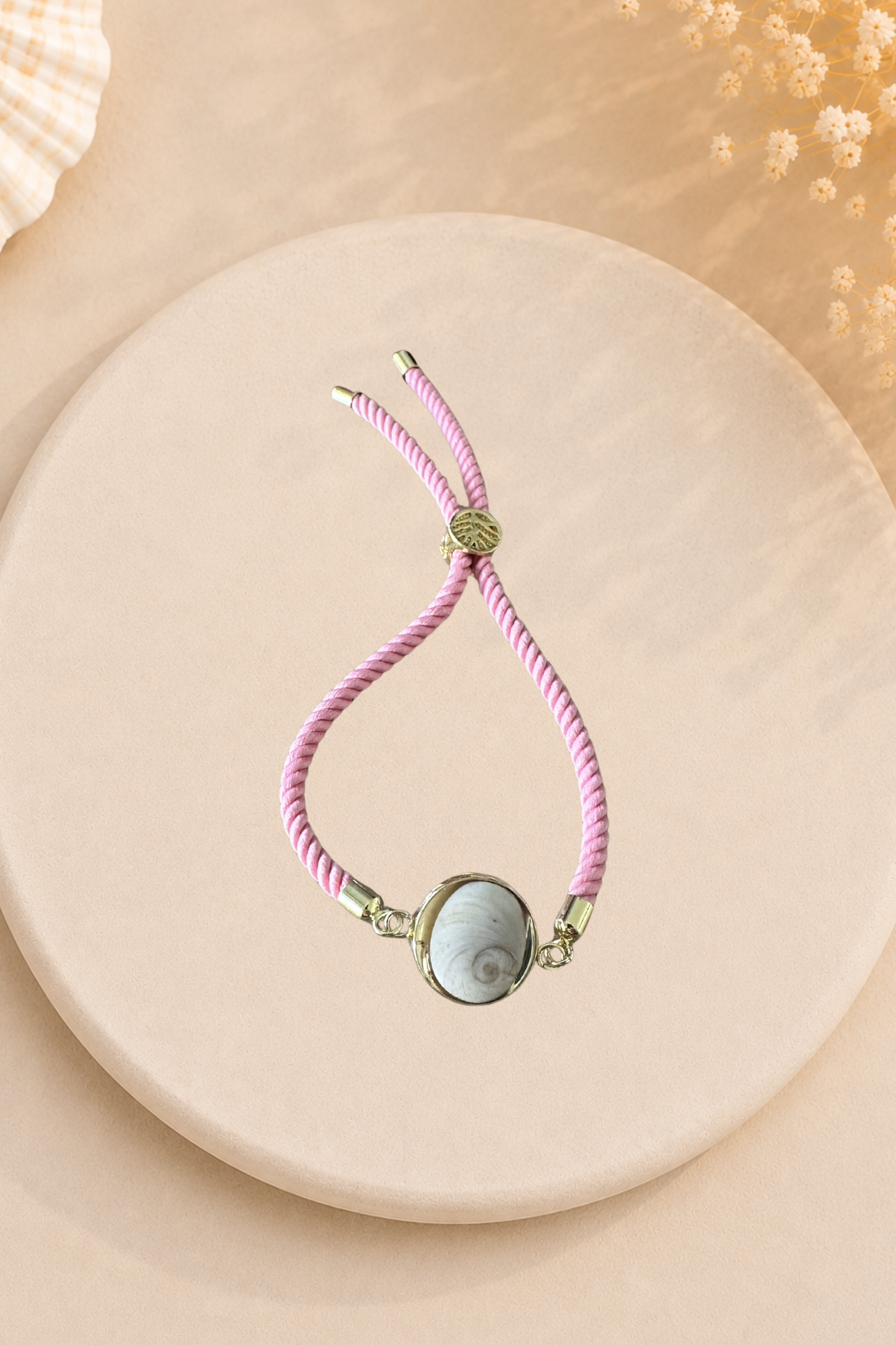 Bracelet femme cordon rose au poignet, médaillon rond doré serti d’un Œil de Sainte‑Lucie blanc, embouts dorés, style chic et marin sur fond gris.
