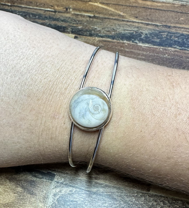 Bracelet jonc argenté avec médaillon rond serti d’un Œil de Sainte‑Lucie blanc, posé sur boîte beige, fond bois, style minimaliste marin.