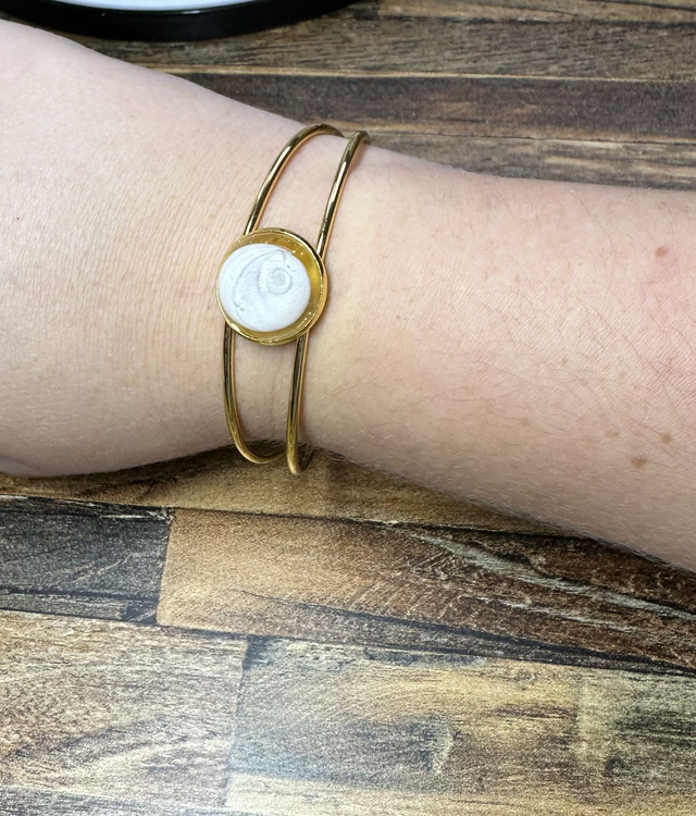Bracelet jonc doré avec médaillon rond serti d’un Œil de Sainte‑Lucie blanc, posé sur boîte beige, fond bois, style minimaliste marin.