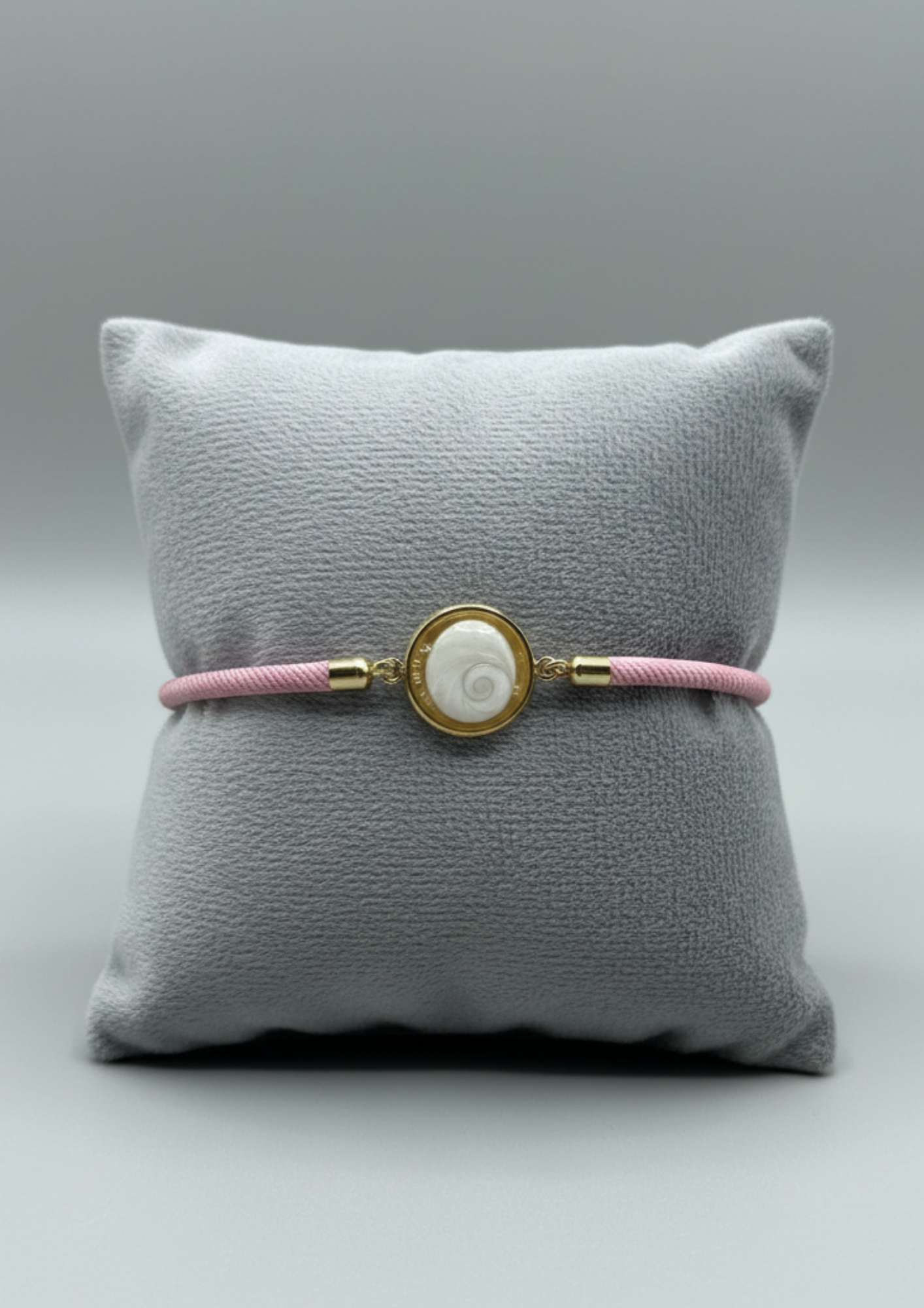 Bracelet femme sur coussin gris, cordon rose fin, médaillon rond doré serti d’un Œil de Sainte‑Lucie blanc, style chic et minimaliste.